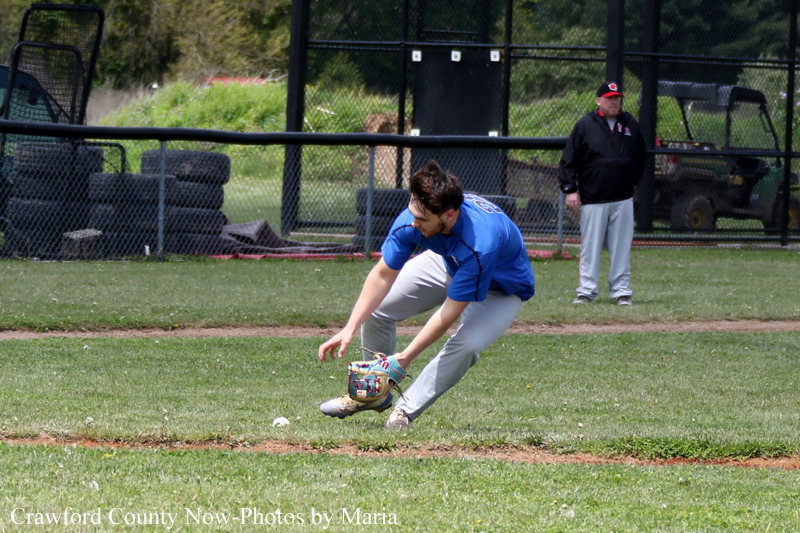 Baseball fielder in a blue shirt and gray pants dives to field a ground ball with his glove near the infield dirt; coach watches from behind the fence.