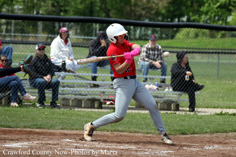 Youth baseball player in a red jersey and white helmet swings a bat, wearing pink gloves, mid-swing on a dirt batter's box.