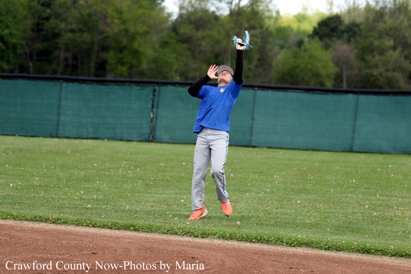 Youth baseball outfielder in a blue jersey catches a high fly ball on a grassy field with a green fence in the background, orange cleats visible.