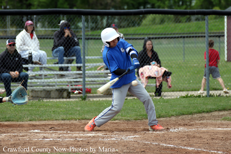 Young baseball batter in a blue uniform swings at a pitch on a dirt field, with a chain-link fence and spectators in the background.