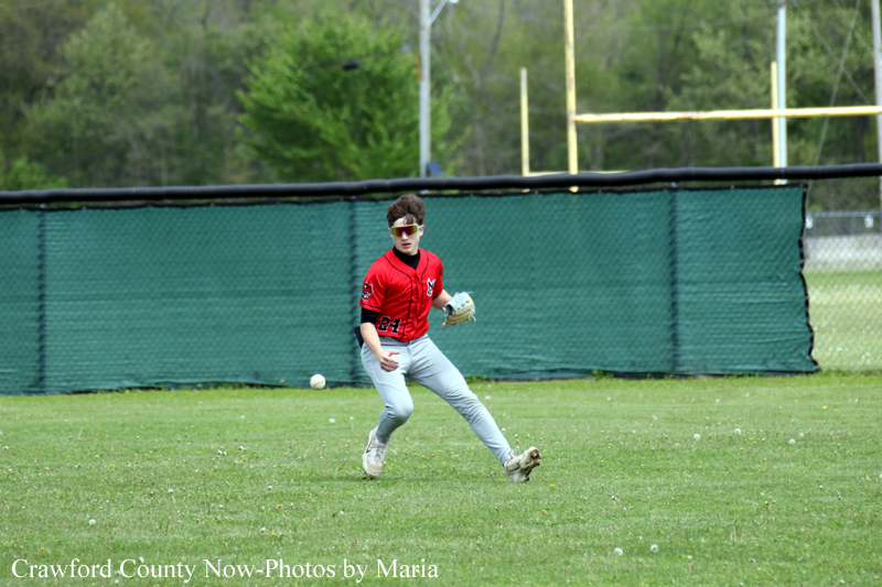 Young baseball fielder in a red jersey and gray pants, glove up, ready to field on a sunny grassy field with a backstop fence behind him.