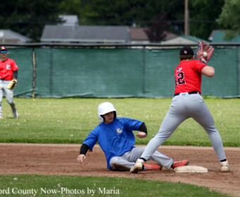 Youth baseball player in blue slides into second base as a red-jersey fielder reaches to tag him; another player watches in the background.