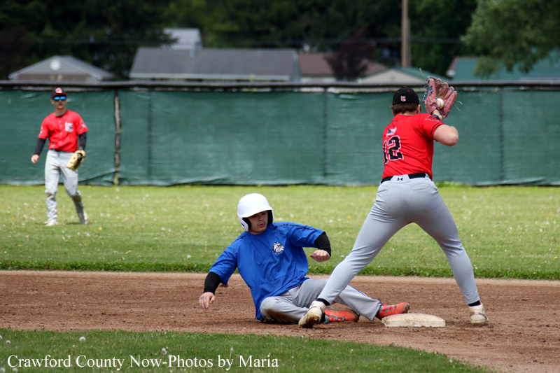 Youth baseball player in blue slides into second base as a red-jersey fielder reaches to tag him; another player watches in the background.