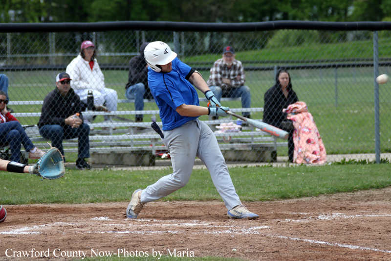Young baseball batter in a blue shirt swings at a pitch during a youth game, with a catcher and fans behind the fence in the background.