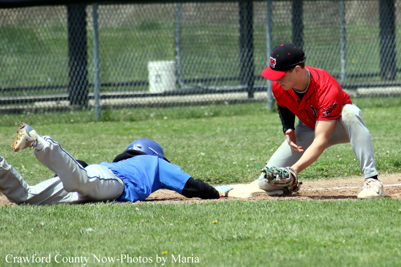 Baseball scene: runner in blue slides into a base as a fielder in red and black reaches to tag him.