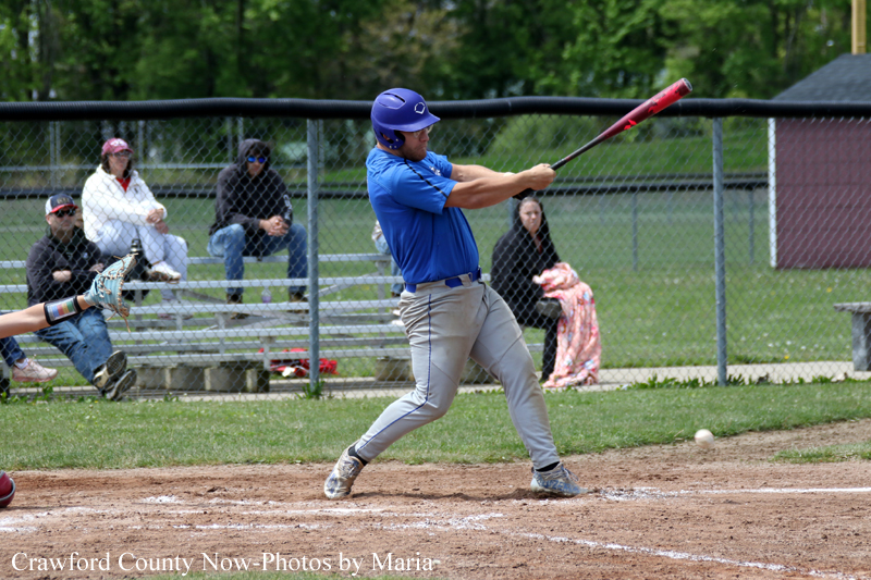 Baseball batter in a blue uniform swings at a pitch during a game, with a catcher and spectators behind the fence in the outfield.