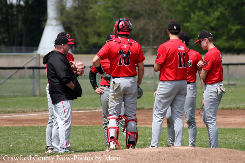 Baseball players in red uniforms huddle on the pitcher's mound with a coach, discussing strategy on a sunny field.