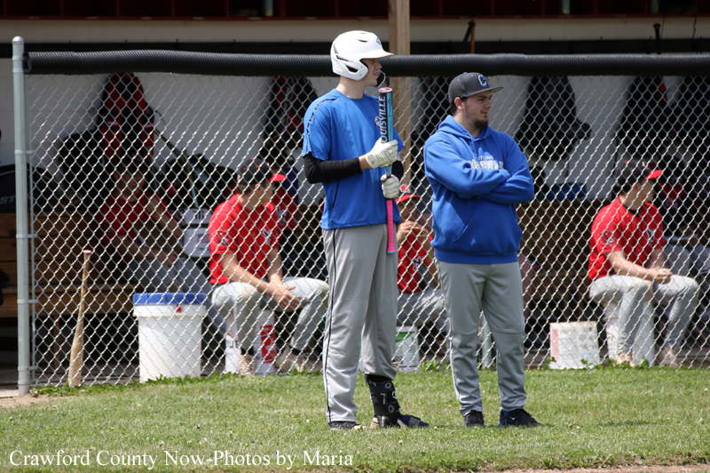 Two baseball players in blue stand by the dugout fence; one wears a white helmet and holds a bat, the other watches with arms crossed.