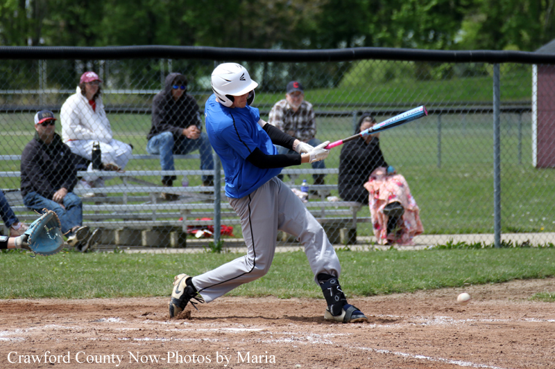Young baseball batter in a blue jersey swings at a pitch at home plate, helmet on, as spectators sit on bleachers behind a chain-link fence.