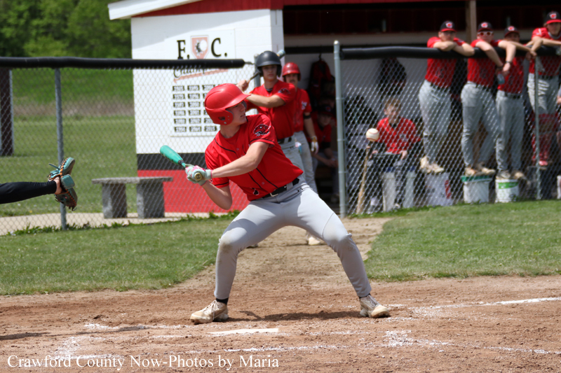 Baseball batter in a red uniform and helmet swings at a pitch at home plate, with a catcher reaching from the left and teammates watching from the dugout behind a chain-link fence.