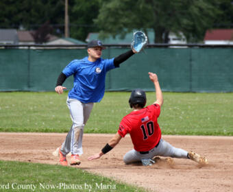 Baseball infielder in blue reaches with his glove as a red-uniform runner slides into a base on a dirt infield.