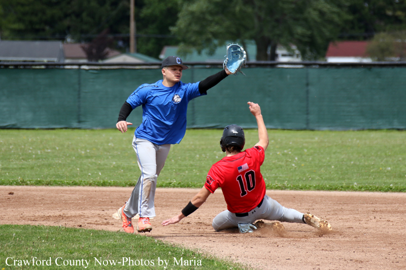 Baseball infielder in blue reaches with his glove as a red-uniform runner slides into a base on a dirt infield.