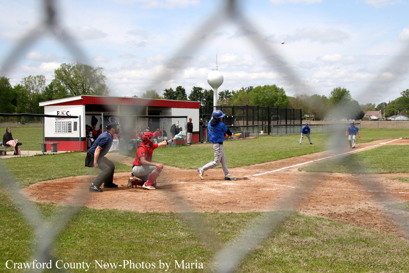 Youth baseball scene: batter in blue swings at home plate as catcher in red crouches and umpire watches nearby. Fence and field in background.