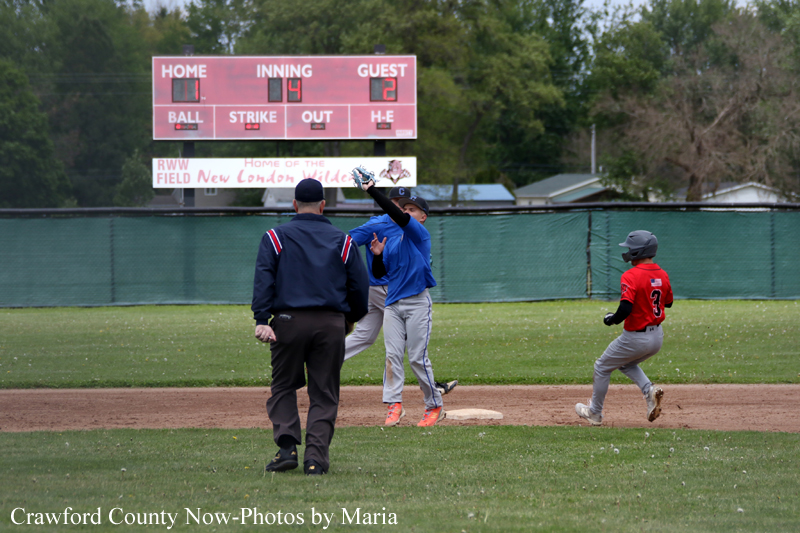 Baseball game in progress with an umpire at home plate as a blue-jersey runner and a red-jersey runner approach first base, scoreboard in the background showing inning 4 and the guest team ahead 2 keys.