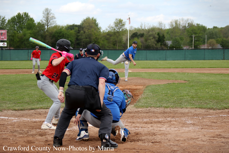 Batter in red swings at home plate as catcher in blue crouches behind him and an umpire stands nearby; pitcher in blue throws from the mound in the background.