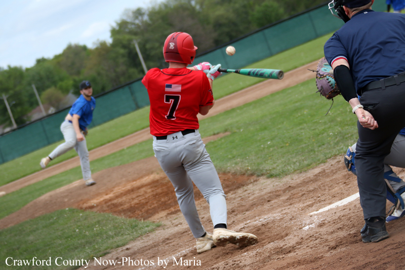 Baseball batter in red jersey with number 7 swings at a pitch, ball in midair near the bat, pitcher in blue in the background and catcher/umpire nearby.