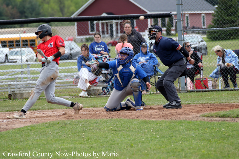 Baseball game at home plate: a runner in red slides toward home as a blue-clad catcher kneels to tag, with the umpire behind and spectators watching from the fence.