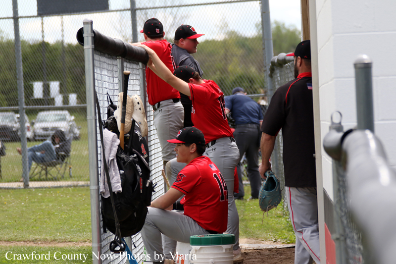 Baseball players in red uniforms sit and stand in a dugout by a chain-link fence, preparing for the game.