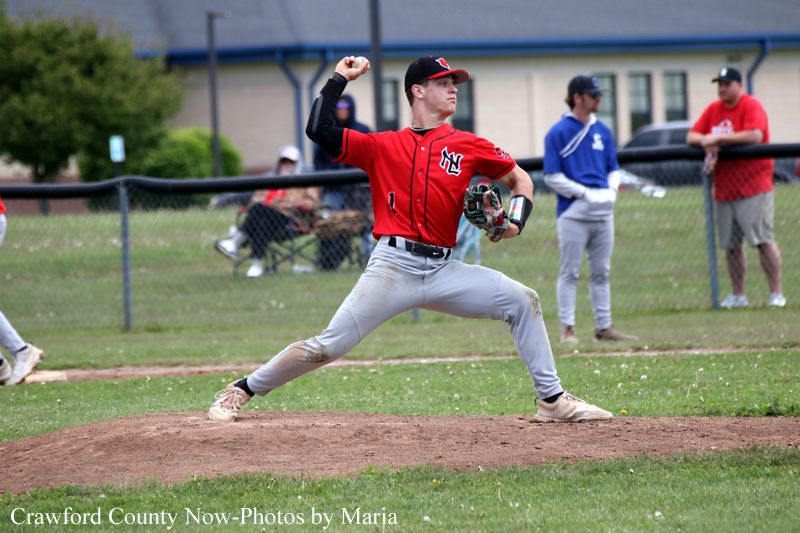 Pitcher in a red uniform delivering a throw from the mound during a baseball game, with teammates and spectators in the background.