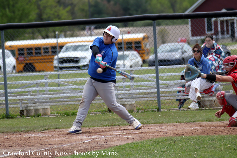 Youth baseball game action: batter in blue swings at a pitch, with a catcher in red gear nearby and a chain-link fence in the background.