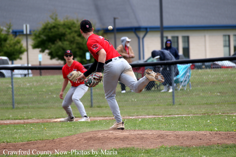 Baseball pitcher in red faces the plate, mid-throw from the mound with a fielder ready in the infield behind him.