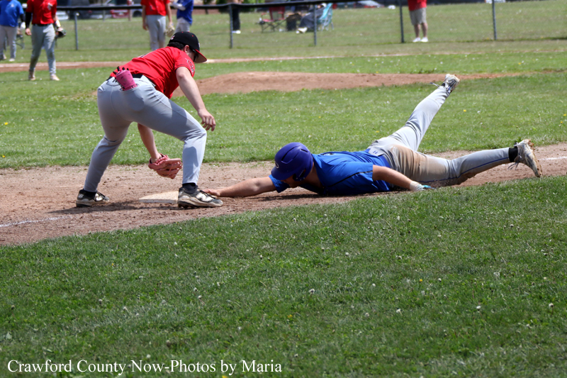 Blue-uniform baserunner slides headfirst into a base as a red-uniform fielder reaches with a glove nearby on a dusty infield.