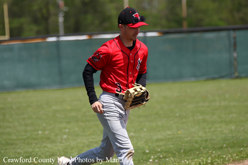 Young baseball player in a red uniform with black undershirt, wearing a cap, walking on the grass with a glove in hand during a game.
