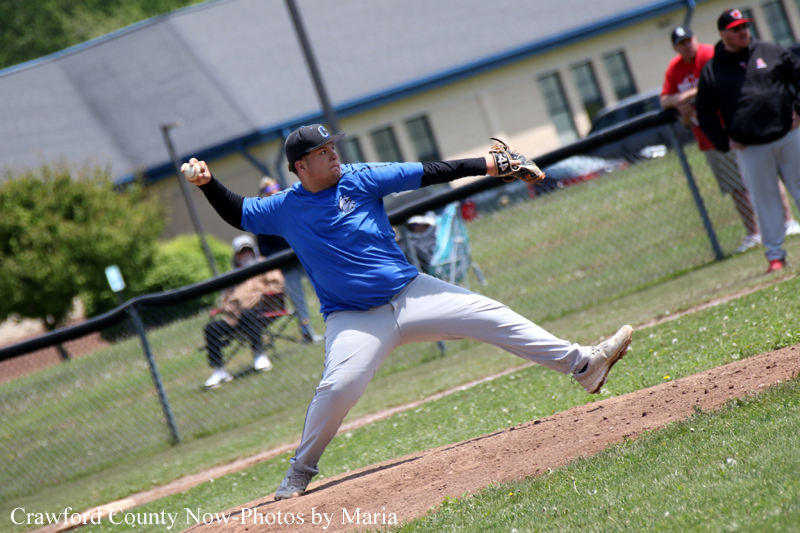 Baseball pitcher in blue jersey on the mound throwing a pitch during a game, with field and spectators in the background