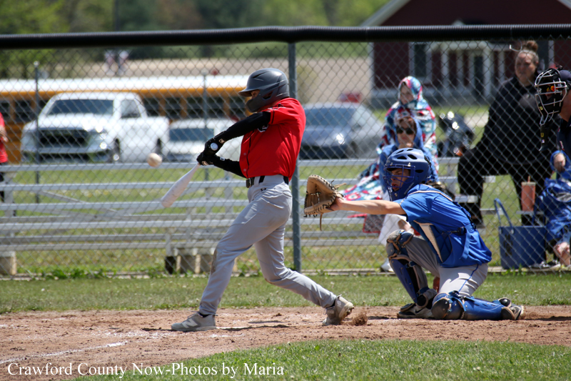 A young batter in a red uniform swings at a pitched ball as the catcher in blue crouches behind home plate on a dusty baseball diamond.