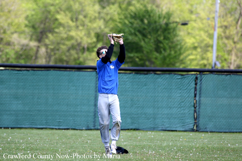 Baseball player in a blue jersey catches a ball overhead on a grassy field with a green fence in the background.