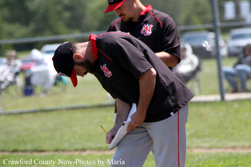 Baseball coach bending over to take notes on a clipboard during a game or practice with another player in the background.