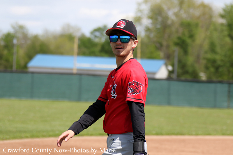Baseball player in a red uniform and cap, wearing dark sunglasses, standing on the infield with a green outfield fence behind him.