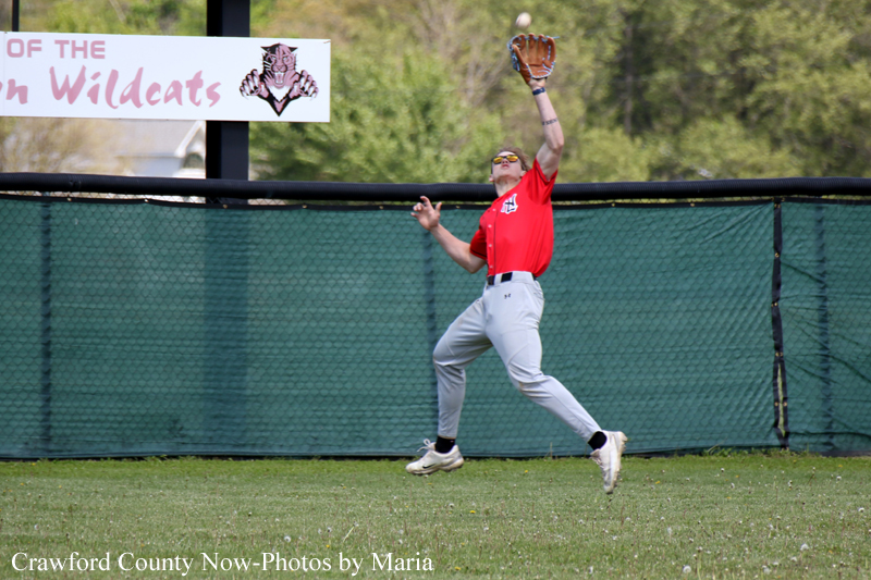Baseball player in a red jersey leaps to catch a ball with a glove near the outfield fence during a game.