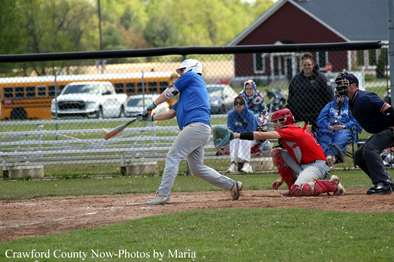 Baseball batter in blue swings at a pitch as the red-gear catcher and umpire crouch behind home plate, with spectators behind a chain-link fence.
