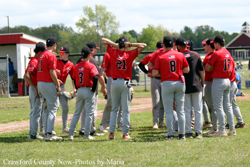 Youth baseball team in red uniforms huddled together on the infield dirt of a sunny field.