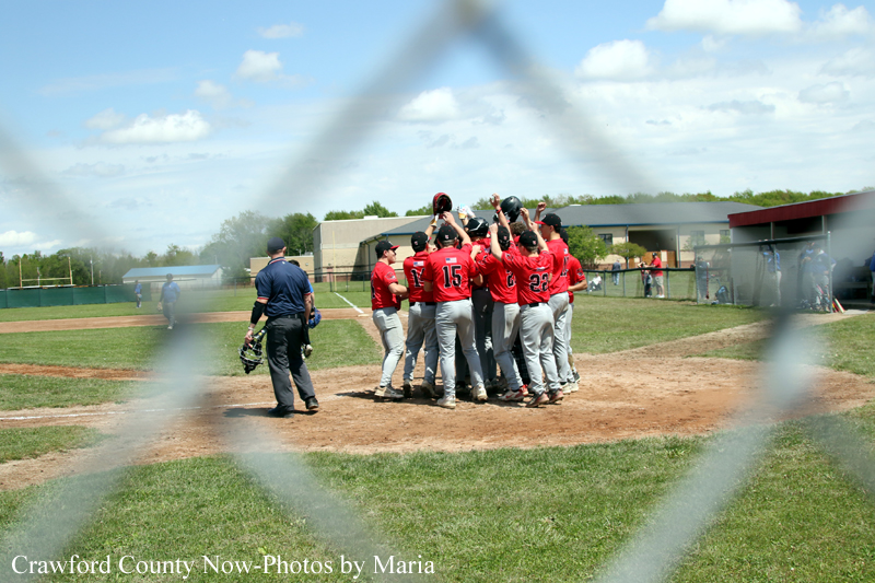 Baseball players in red jerseys huddle on the infield, seen through a chain-link fence with a clear blue sky behind them, celebrating a moment on the field.