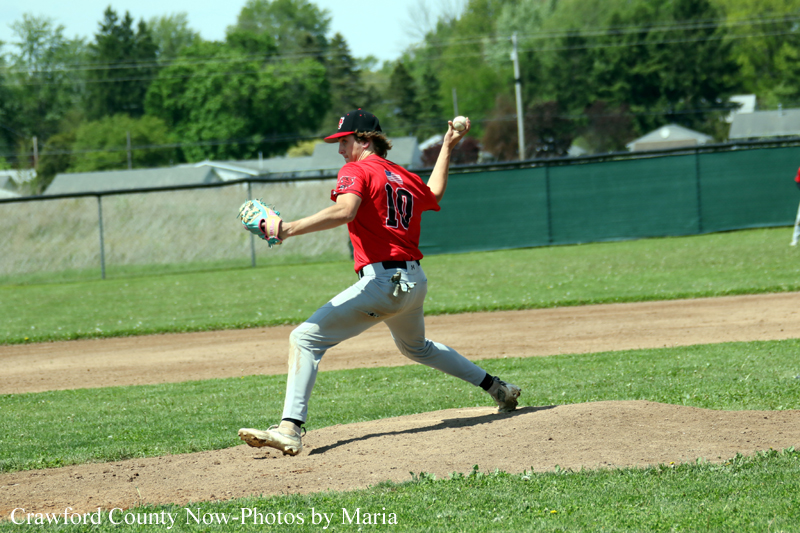 Baseball pitcher in a red uniform throws a pitch from the mound on a sunny field with green grass and a fence in the background.