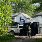 Soldier in camouflage stands next to a black armored vehicle in a residential driveway, with white houses and trees nearby.