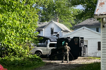 Soldier in camouflage stands next to a black armored vehicle in a residential driveway, with white houses and trees nearby.