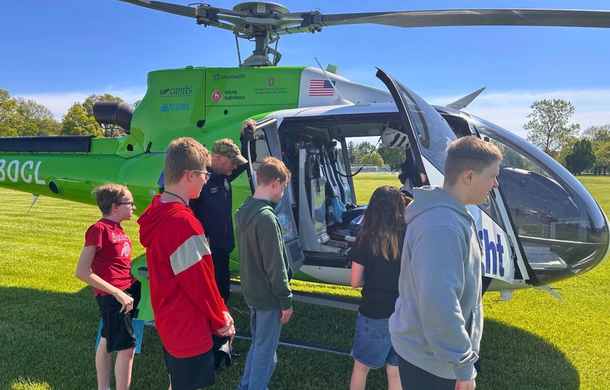 Kids and a guide boarding a bright green helicopter on a sunny grassy field.