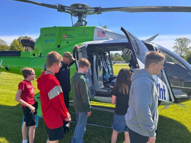 Kids and a guide boarding a bright green helicopter on a sunny grassy field.