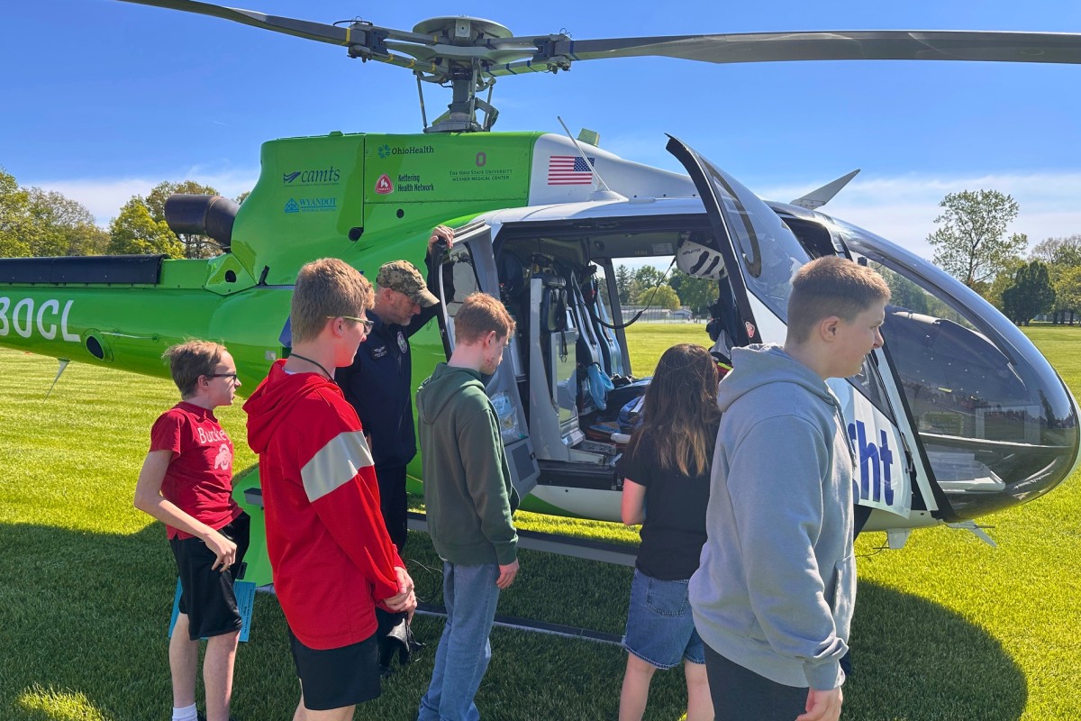 Kids and a guide boarding a bright green helicopter on a sunny grassy field.