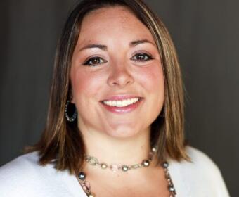 Smiling woman with shoulder-length brown hair wearing a beaded necklace and hoop earrings.