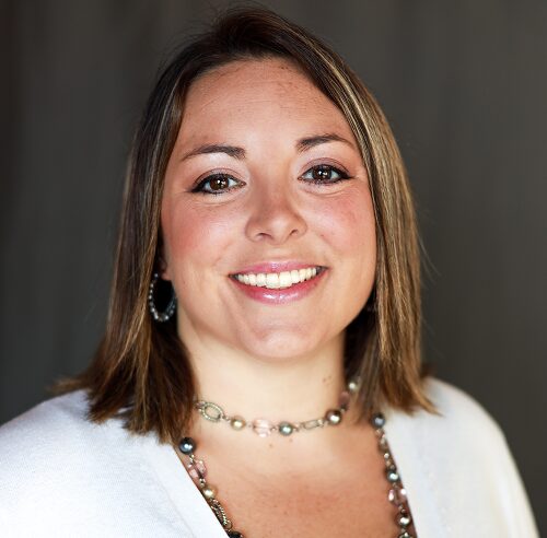 Smiling woman with shoulder-length brown hair wearing a beaded necklace and hoop earrings.