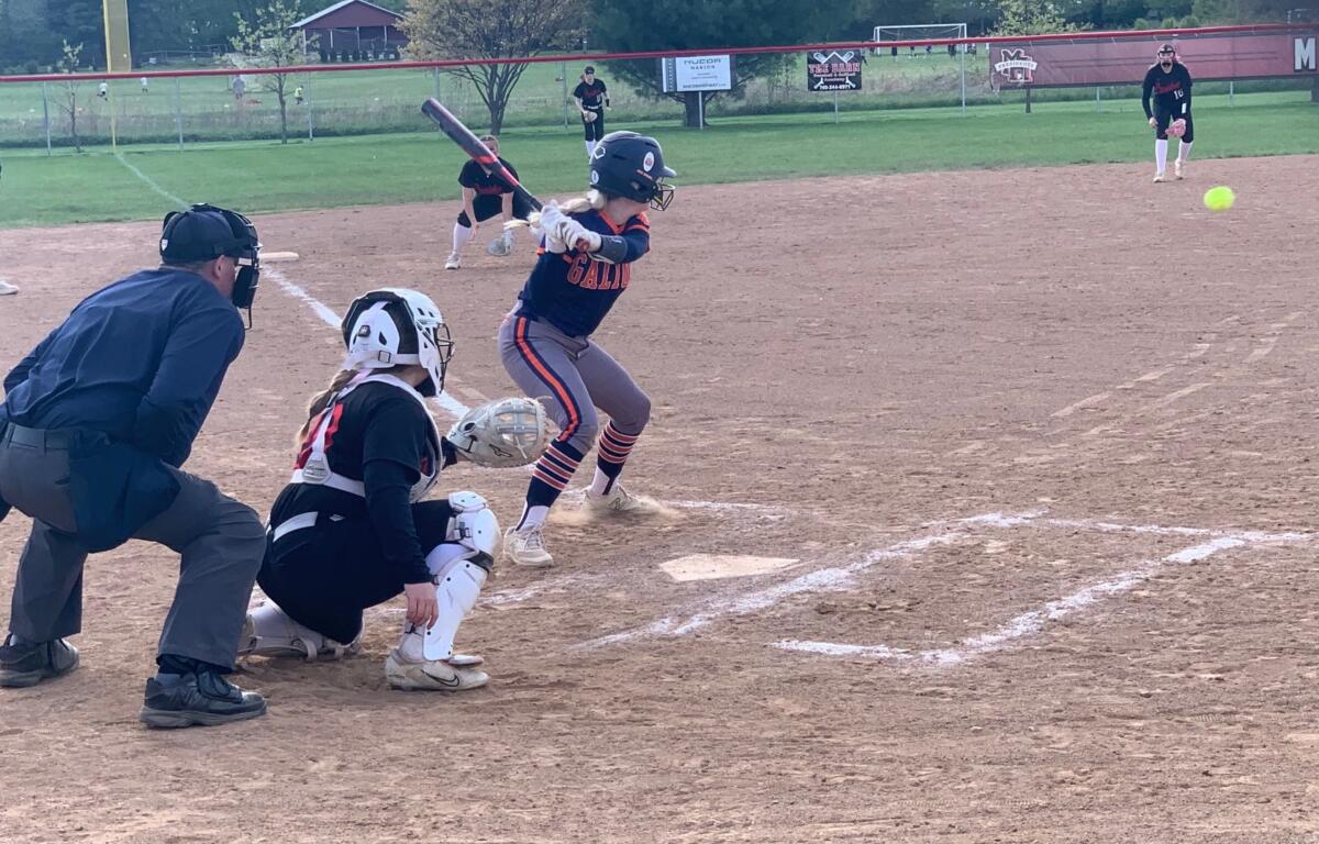 Young batter swings at a pitch in a youth softball game, with catcher and umpire behind home plate on the dirt field.