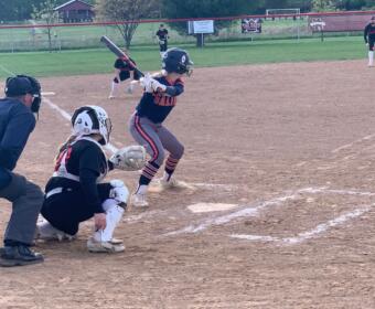 Young batter swings at a pitch in a youth softball game, with catcher and umpire behind home plate on the dirt field.