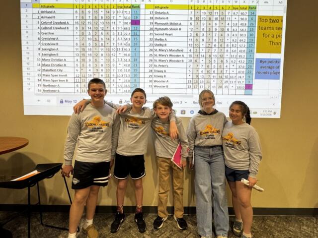 Five children in gray Academic Challenge shirts pose with arms around each other in front of a projected spreadsheet on a yellow wall.