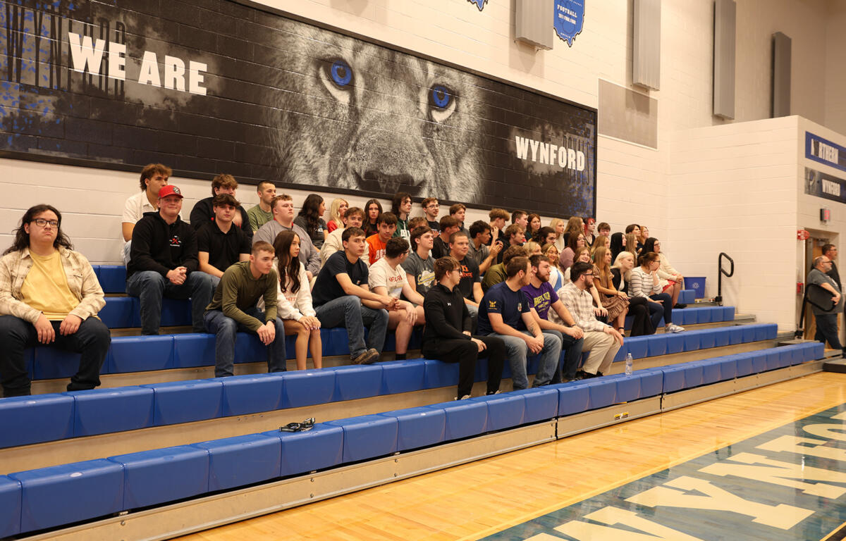 A large group of students sitting on blue gym bleachers in a school gym, posing for a group photo under a wall mural that reads 'WE ARE WYND' and features a lion's eyes close-up.