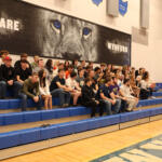 A large group of students sitting on blue gym bleachers in a school gym, posing for a group photo under a wall mural that reads 'WE ARE WYND' and features a lion's eyes close-up.