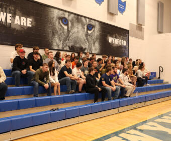 A large group of students sitting on blue gym bleachers in a school gym, posing for a group photo under a wall mural that reads 'WE ARE WYND' and features a lion's eyes close-up.